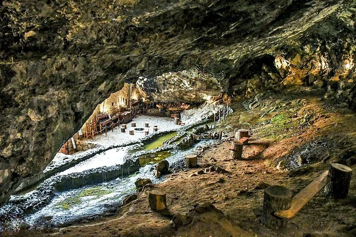 Inside a large natural cave with a stream and wooden seating areas.